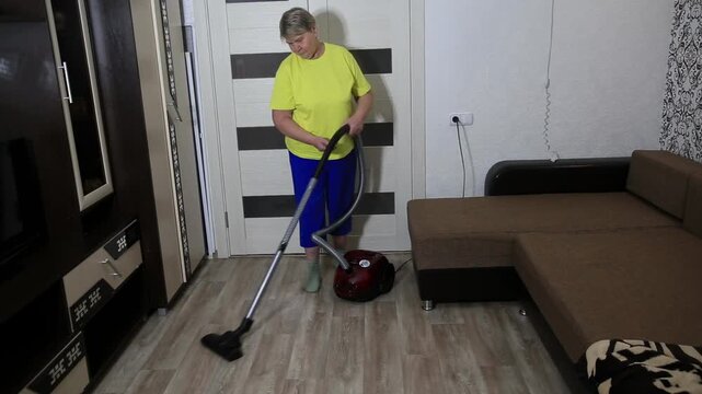Adult woman performing household chore of cleaning floor with red vacuum cleaner in bright room of home, diligently vacuuming during day, maintaining cleanliness.