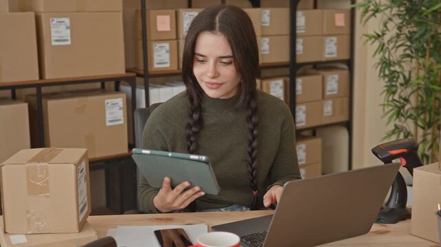 Woman holding tablet and typing on laptop in a building shipping area, surrounded by packages and boxes; entrepreneurship focused.