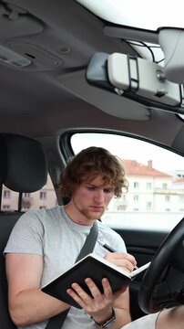 Vertical screen focused learner records answers in his notebook from the driver seat of a parked car while reviewing road regulations for an upcoming driving license test
