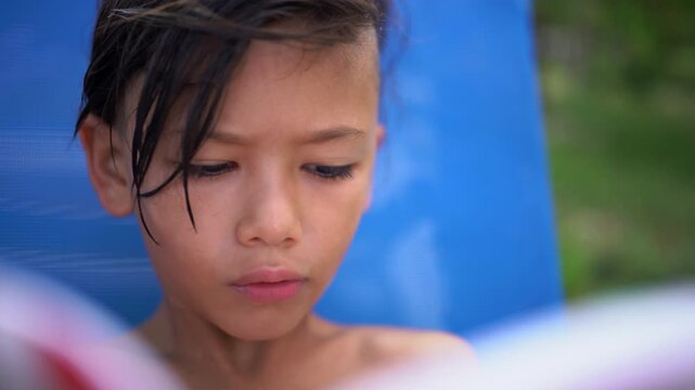 A close-up of a boy with wet hair reading a comic book beside the swimming pool