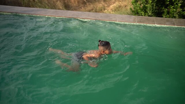 A boy swims across the pool to the edge, spits water from his mouth, then looks at the camera and laughs, High Angle Shot