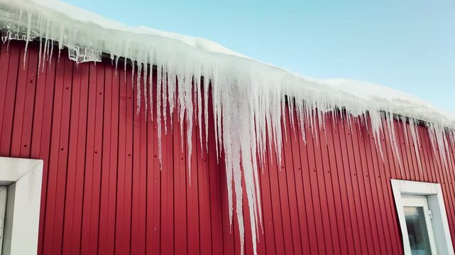Red building exterior with snow accumulation on roof and numerous long, sharp icicles hanging from eaves, clear blue sky visible above, scene portrays a bright, cold winter day.