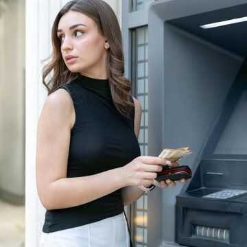Woman looking cautiously around while holding cash near an ATM, conveying concern about personal safety and security during a financial transaction.