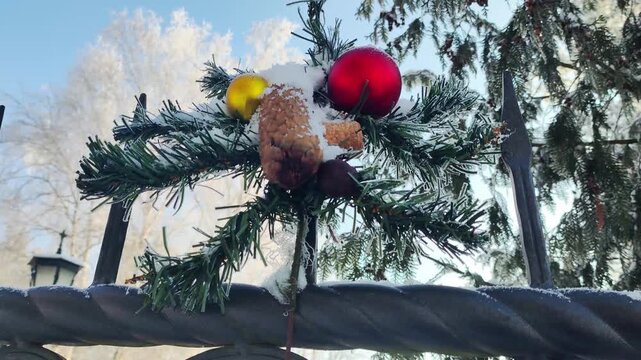 Christmas decoration of artificial pine branches, red and yellow ornaments, pinecone, and fresh white snow on top of metal fence with frosted trees under clear blue sky of winter day