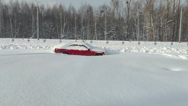 Red car covered in deep white snow, abandoned on roadside next to line of bare winter trees under cloudy sky, illustrating harsh weather conditions and vehicle immobilization