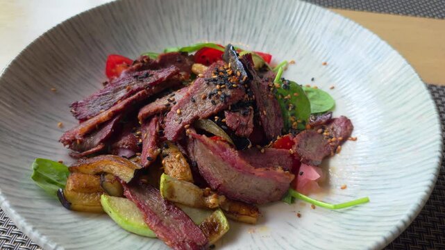 Gourmet dish featuring thinly sliced grilled beef, fresh spinach, red tomatoes, roasted eggplant, and zucchini, garnished with black and white sesame seeds, presented in decorative bowl.