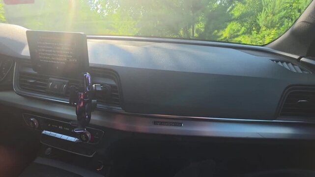 Interior view of car dashboard on sunny day, bright sunlight illuminates cockpit, vehicle travels rural road through lush green forest with infotainment screen displaying information