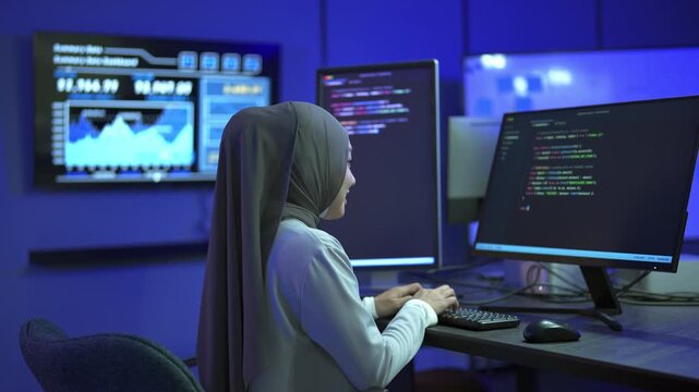 Young Muslim woman in hijab typing programming code on multiple computer monitors in a modern office.