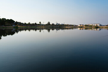 Obraz premium Calm water surface reflecting distant village buildings under clear sky.