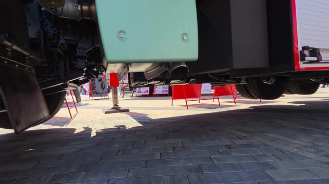 Underside view of heavy duty vehicle chassis with hydraulic support system extended on paved outdoor surface, highlighting suspension components and exhaust pipes during daytime exhibition