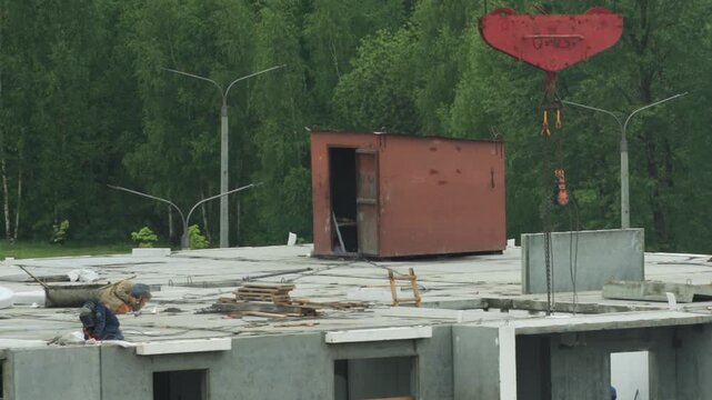 Worker welding on concrete roof of panel house construction site with red crane hook and temporary shed against green forest background.