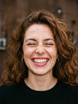 Young woman laughing with eyes closed in outdoor rustic setting, freckled face and curly hair framed by wooden fence background conveying joyful candid portrait