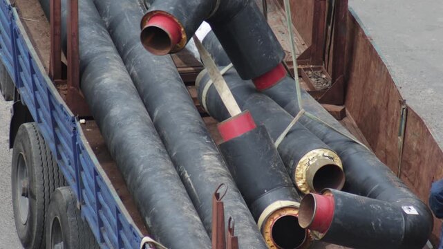 Unloading insulated pipeline sections from flatbed truck bed transporting construction material. Large black pipes with red caps and protective insulation move from vehicle.