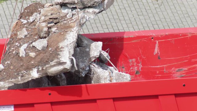 Loading concrete debris construction waste into red dump truck bed with rebar visible during demolition project day outdoors, showing heavy lifting process