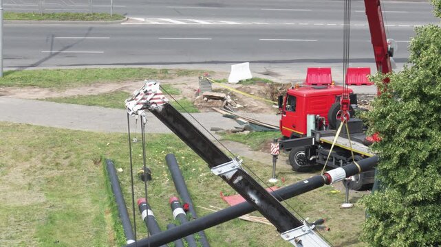 Red truck crane lifting large black pipes at construction site alongside empty road, showing infrastructure development and heavy machinery work during day outdoors.