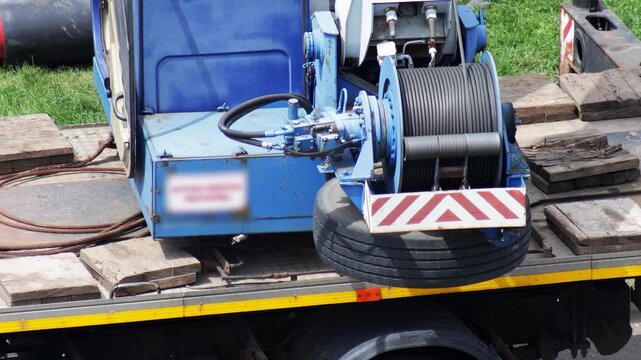 Blue truck-mounted winch system with cable spool and hydraulic mechanisms on flatbed truck, industrial heavy lifting equipment, construction machinery detail, slow motion