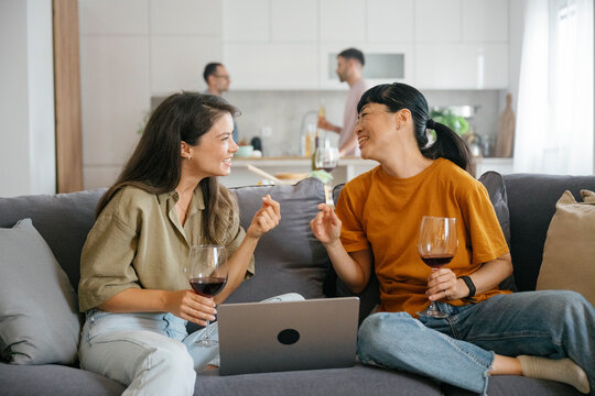 Friends laughing and talking on couch with wine and laptop at home