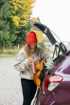 Woman packs her backpack at the car trunk in an autumn forest