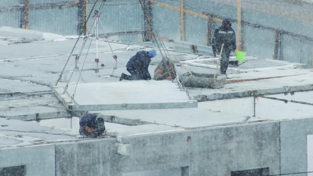 Construction workers labor on building site roof during heavy winter snowfall, placing concrete slabs with crane rigging system, enduring challenging weather conditions
