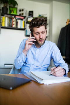 A man writes notes while calling on a smartphone in his home office