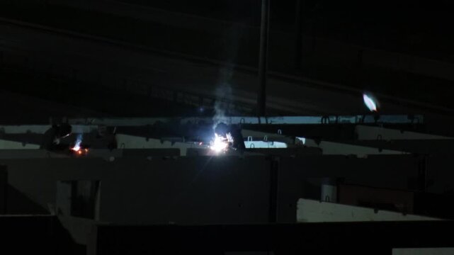 Night construction scene showing worker performing welding operation bright sparks smoke rising from metal connection concrete walls surrounding workspace