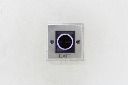 No-touch exit button with illuminated purple ring mounted on stainless steel plate beside worn white wall in close-up view of public building access control hardware