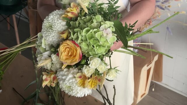 Woman florist in apron holds bouquet with roses, chrysanthemums and hydrangeas at workshop table. Small business floristry, handmade bouquet craft, flower shop workflow, artisan entrepreneurship