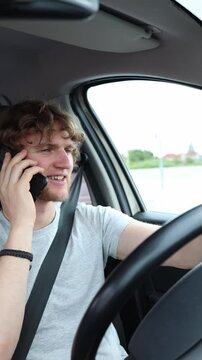 Vertical screen relaxed driver smiles during a cheerful phone conversation behind the wheel while cruising comfortably through urban traffic in his carefree positive mood