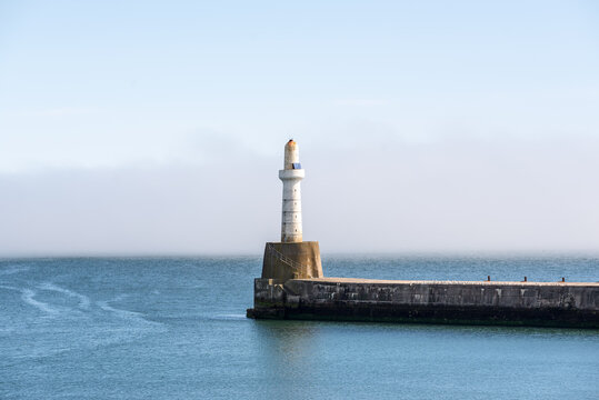 A haar is rolling in. Historic Aberdeen Harbour lighthouse standing on the breakwater, surrounded by calm blue sea and misty coastal scenery on a summer day.