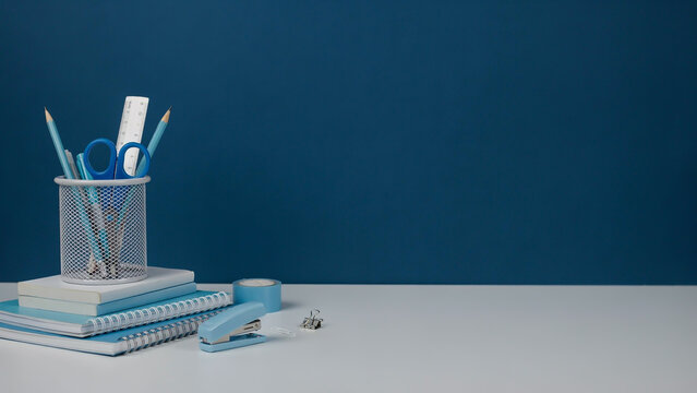 A minimalist still life photograph of office supplies, including a white mesh pen holder, a blue spiral notebook, a stapler, and a roll of tape