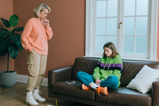 Caucasian middle aged woman standing near sad teenager girl sitting on sofa and avoiding eye contact, showing family conflict or parenting stress. For counseling, mental health, parenting support