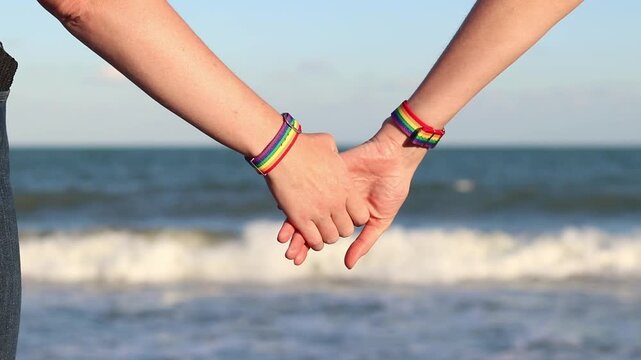 Same-sex couple with LGBTQ+ bracelets on their wrists. Two girls holding hands against the sea. Couple holding hands on the beach, showing off their pride bracelets. Love and equality concept