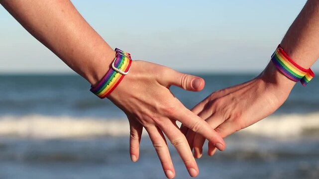 Same-sex couple with LGBTQ+ bracelets on their wrists. Two girls holding hands against the sea. Couple holding hands on the beach, showing off their pride bracelets. Love and equality concept
