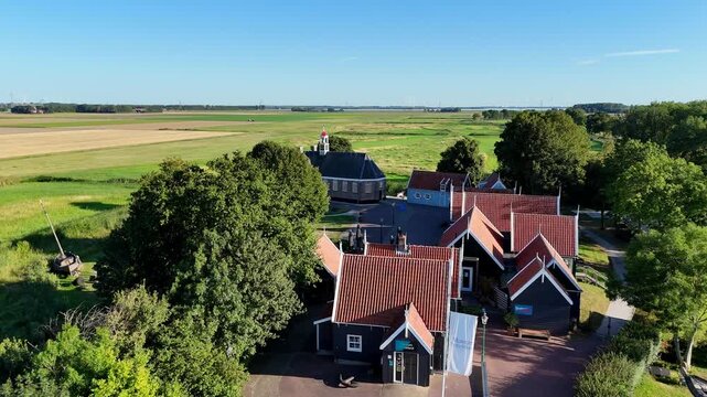 A scenic drone shot capturing the historic former island of Schokland in the Netherlands on a bright, sunny day. Flying over the entrance towards the church, revealing the cannon. Single traveler.
