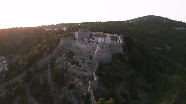 Hvar Fortress crowns a forested hill above the town and harbor, as a drone glides past at golden hour. Boats fill the bay and nearby islands.
