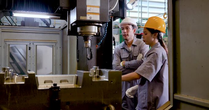 Asian woman technician stands near CNC fixture as Asian man engineer explains status, guiding inspection, machine check, and data driven coordination for efficient manufacturing operation inside plant