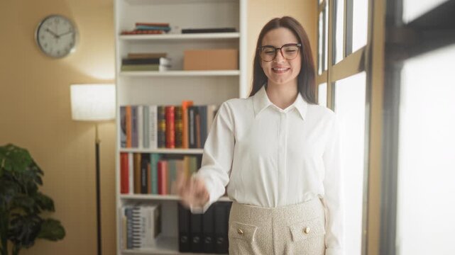 Woman adjusting glasses with hand, smiling and wearing white blouse and tweed skirt in office by window and bookshelf; warm confidence.