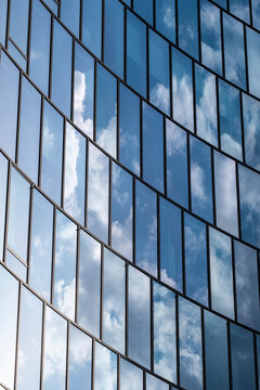 Glass facade of modern architecture with blue windows reflecting clouds and sky across a geometric exterior pattern of repeating rectangular panels