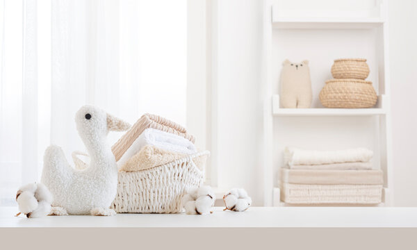 Terry towels in white basket, cotton fluffy flowers and soft toy duck on white table close up with copy space over curtained window and bathroom shelf blur background