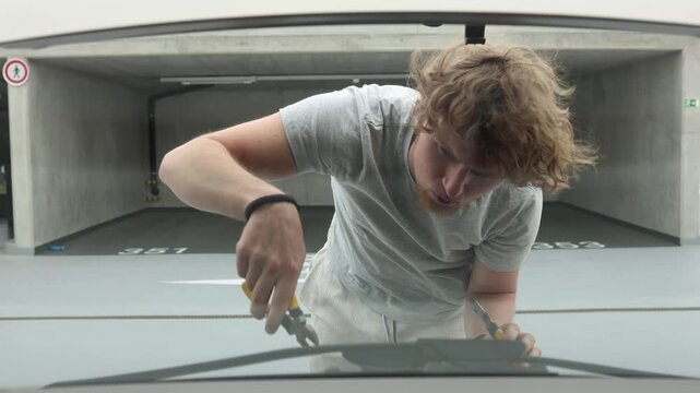 Young car owner repairs his vehicle under the open hood in a parking garage using hand tools while independently fixing the engine problem without professional assistance