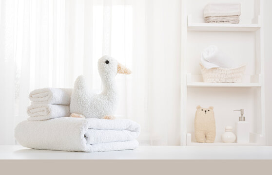 White towels stack and soft toy duck on white table with design space over curtained window and spa bathroom shelf blur background