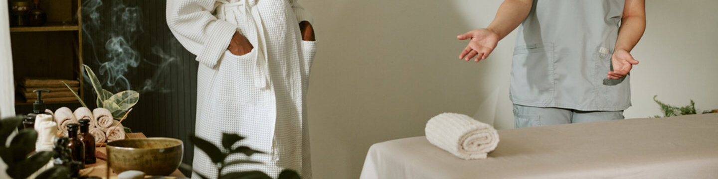 Header of female masseuse welcoming young adult African American woman in bathrobe to massage table. Image represents wellness services and is ideal for spa marketing