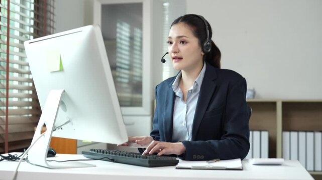 Professional call center agent wearing headset providing friendly customer service and typing on keyboard while working at her desk in the corporate telemarketing office