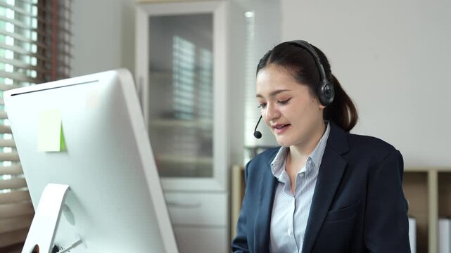 Professional call center agent providing customer support, smiling Asian operator wearing a headset with microphone and typing on a computer keyboard in a bright office