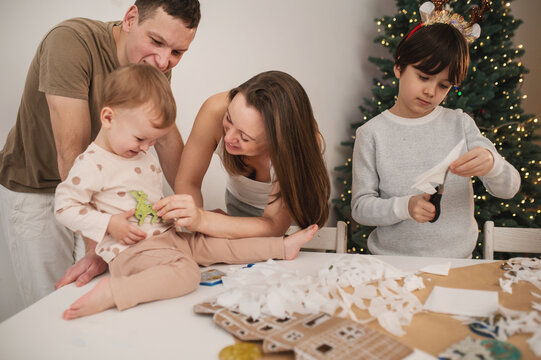 Children and parents celebrate Christmas through creative crafting, making handmade paper decorations together while surrounded by festive lights and cozy seasonal home atmosphere.