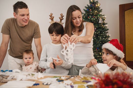 Happy family cut paper snowflakes near Christmas tree, celebrating Christmas with together. New Year's preparations and decorations. Paper crafts. 