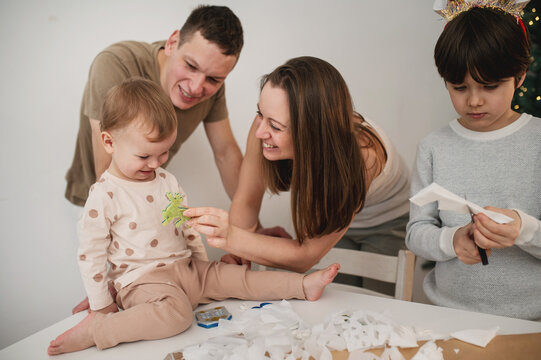 Happy family gathers around table for Christmas crafts as children create handmade decorations, enjoying warm holiday moments together beside glowing festive Christmas tree.