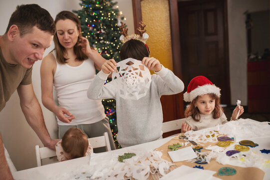 Happy family cut paper snowflakes near Christmas tree, celebrating Christmas with together. New Year's preparations and decorations. Paper crafts. 