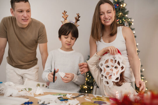 Happy family cut paper snowflakes near Christmas tree, celebrating Christmas with together. New Year's preparations and decorations. Paper crafts. 