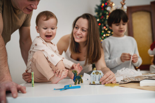 Family celebrates Christmas together as parents and children craft festive paper decorations at table beside glowing Christmas tree, enjoying creative holiday time indoors.